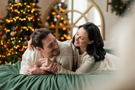 Porträt eines fröhlichen Paares, das zusammen auf dem Bett in der Nähe des geschmückten hellen Weihnachtsbaums mit Lichtern liegt