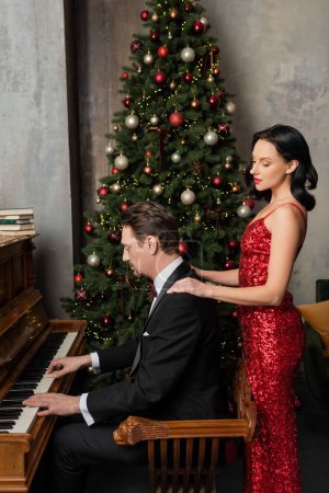 Wohlhabendes Paar, brünette Frau in rotem Kleid, neben Ehemann stehend, der am Klavier spielt, Frohe Weihnachten