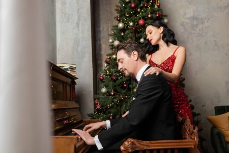 Reiches Paar, brünette Frau in rotem Kleid, neben Ehemann stehend, der am Klavier spielt, Frohe Weihnachten