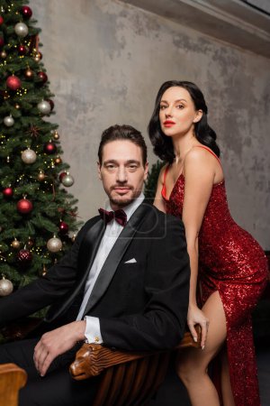 rich couple, pretty woman in red dress standing near husband next to decorated Christmas tree