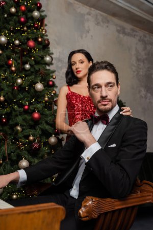 Wohlhabendes Paar, hübsche Frau in rotem Kleid neben Ehemann neben geschmücktem Weihnachtsbaum