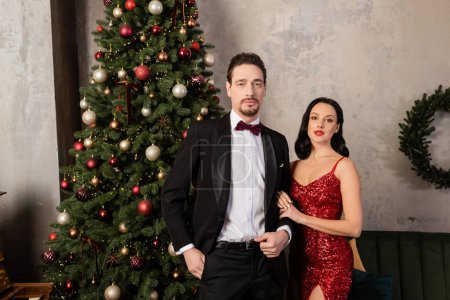 pareja rica, mujer elegante en vestido rojo de pie cerca del hombre en esmoquin y árbol de Navidad decorado