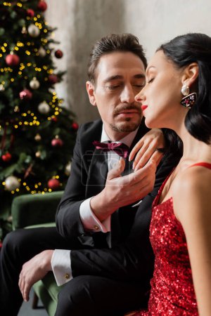 gentleman in tuxedo gently touching face of attractive woman with brunette hair near Christmas tree
