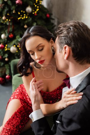 gentleman in suit sitting on sofa and kissing cheek of woman in red dress near Christmas tree