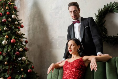 hombre con esmoquin de pie detrás de una hermosa mujer vestida de rojo cerca del árbol de Navidad, gente rica