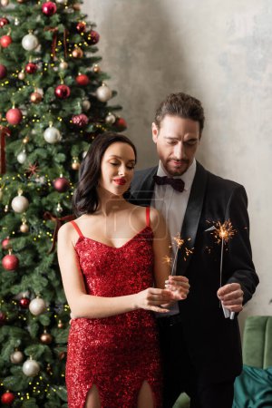 hermosa pareja rica en traje formal y elegante celebración de brillantes chispas cerca del árbol de Navidad