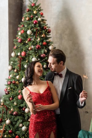 beautiful wealthy couple in formal attire smiling and holding bright sparklers near Christmas tree