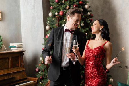 pareja alegre rica en atuendo formal sosteniendo copas de champán y bengalas cerca del árbol de Navidad