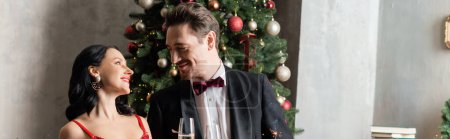 wealthy and happy couple in formal attire holding champagne glasses near Christmas tree, banner