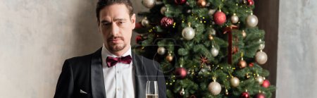 Reicher Mann im Smoking mit Fliege hält Champagnerglas neben geschmücktem Weihnachtsbaum, Banner