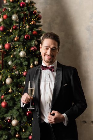 glücklicher reicher Mann im Smoking mit Fliege hält Champagnerglas neben geschmücktem Weihnachtsbaum
