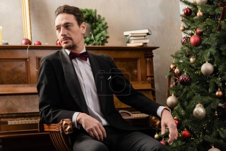 eleganter Herr in formeller Kleidung mit Fliege am Klavier sitzend und geschmücktem Weihnachtsbaum