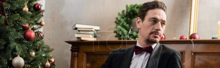 elegant gentleman in tuxedo with bow tie sitting near piano and decorated Christmas tree, banner