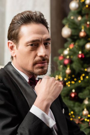 portrait rapproché de gentleman élégant en smoking avec noeud papillon regardant la caméra près de l'arbre de Noël