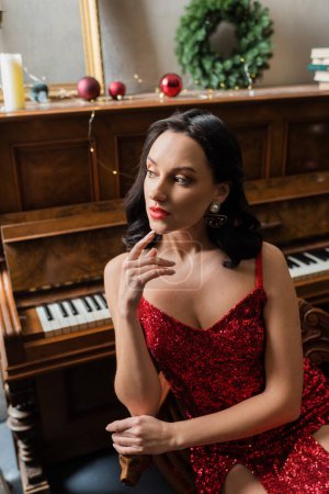 mujer atractiva en vestido rojo elegante sentado cerca del piano y la corona de Navidad, la vida rica