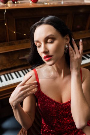 kultivierte Frau mit geschlossenen Augen, die im eleganten roten Kleid neben dem Klavier sitzt, wohlhabendes Leben