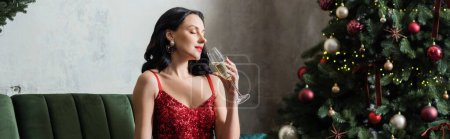 beautiful woman in red dress enjoying taste of champagne near decorated Christmas tree, banner