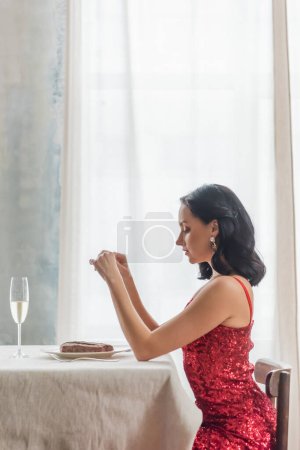 Téléchargez les photos : Vue latérale de jolie femme en robe rouge assise à table avec verre de champagne et steak - en image libre de droit