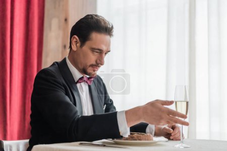 hombre guapo en esmoquin sentado en la mesa de comedor con copa de champán y carne de res en el plato