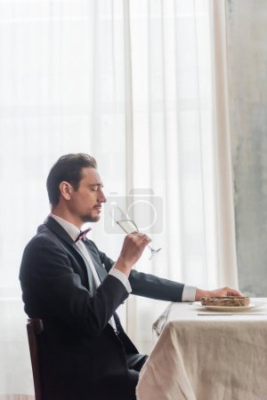 handsome man in tuxedo enjoying taste of champagne and sitting at table with beef steak on plate