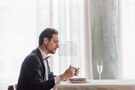 schöner Mann im Smoking genießt Geschmack von Rindersteak auf Teller in der Nähe von Champagner im Glas auf dem Esstisch