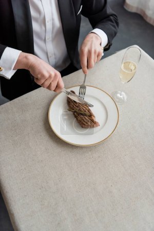 Photo for Top view of wealthy man in tuxedo cutting delicious beef steak on plate near glass of champagne - Royalty Free Image