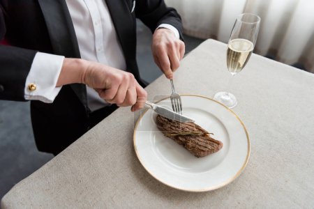 Overhead-Ansicht des reichen Mannes im Smoking schneidet köstliche Rindfleisch-Steak auf dem Teller in der Nähe von Glas Champagner