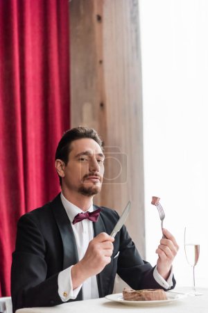 wealthy gentleman in tuxedo enjoying taste of beef steak near champagne in glass on dining table