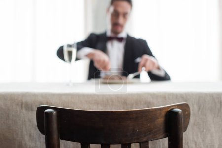 focus on wooden chair, wealthy gentleman in tuxedo enjoying dinner at home, natural light