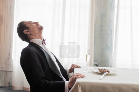Herr im Smoking fühlt sich satt nach leckerem Essen am Tisch, Rindersteak und einem Glas Champagner
