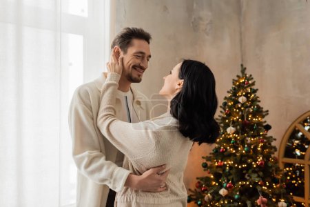 freudiges Paar in bequemer Heimkleidung lächelt und umarmt neben geschmücktem Weihnachtsbaum im Schlafzimmer