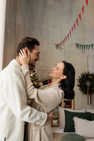 mujer alegre en el hogar cómodo desgaste abrazando marido cerca del árbol de Navidad en el dormitorio, alegría