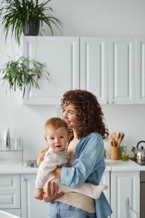 Foto de Mujer alegre con el pelo ondulado abrazando hija pequeña en la cocina en casa, el amor y la ternura - Imagen libre de derechos