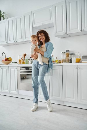 full length of smiling woman with little daughter looking at camera in kitchen with white interior