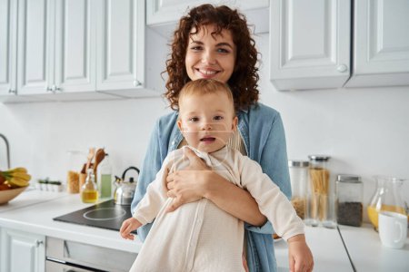 liebevolle Mutter mit welligem Haar umarmt entzückende Tochter im Strampler zu Hause in der Küche, Familienglück