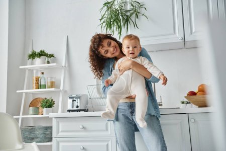 überglückliche Frau hält kleine Tochter im Strampler in Küche mit zeitgenössischem Interieur, gemütlichem Zuhause
