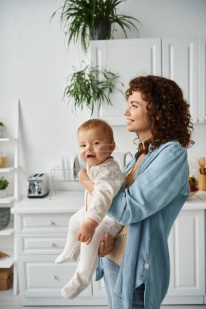 cheerful child in romper looking at camera in hands of mother in kitchen at home, happy childhood