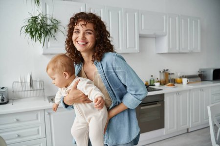 Foto de Mujer excitada con el pelo ondulado sosteniendo niño pequeño y sonriendo a la cámara en la acogedora cocina en casa - Imagen libre de derechos