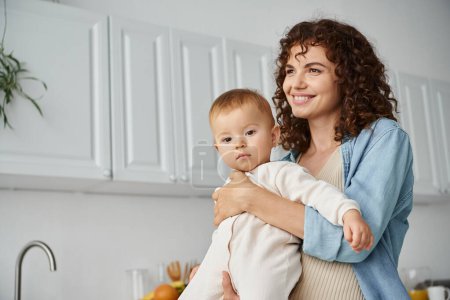 Frau mit lockigem Haar und glücklichem Lächeln, die in der Nähe von Kleinkindtochter in Küche wegsieht, Mutterschaft
