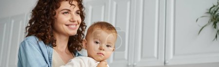 Photo for Happy woman with wavy hair smiling and looking away near adorable baby girl in kitchen, banner - Royalty Free Image