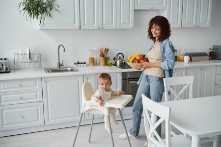 smiling woman with bowl of fresh fruits near child in baby chair in modern kitchen, morning mealtime