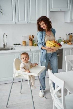 joyful woman holding bowl of fresh fruits near kid in baby chair in contemporary kitchen, breakfast