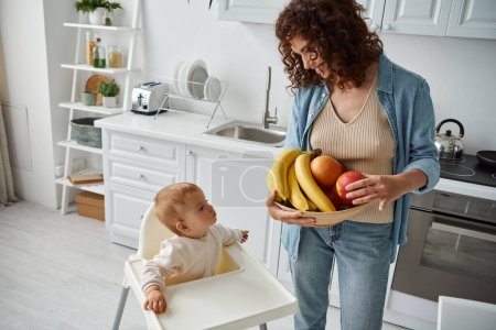 mujer feliz con tazón de plátanos y manzanas cerca de la pequeña hija sentada g en silla de bebé en la cocina