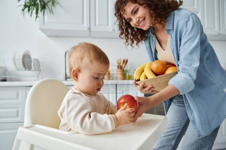 cheerful woman giving ripe apple to toddler daughter sitting in baby chair in kitchen, breakfast