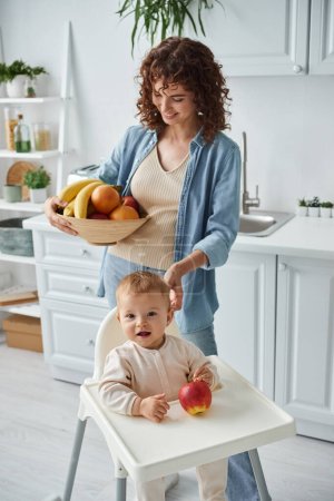 niño emocionado sentado en silla de bebé cerca de manzana madura y mirando a la cámara cerca de mamá con frutas frescas