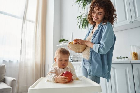 joyful woman with bowl of fresh fruits looking at toddler daughter holding ripe apple in baby chair