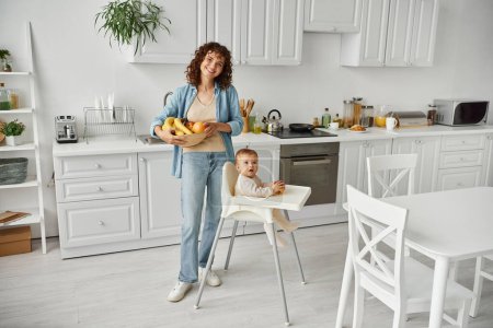 pleased woman with fresh fruits looking at camera near toddler child in baby chair in modern kitchen