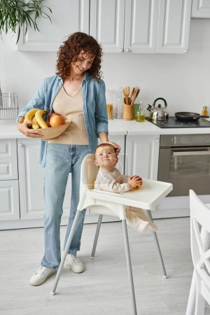 mujer sonriente con frutas frescas acariciando la cabeza de niña sentada en silla de bebé, mañana en la cocina