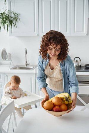 mujer rizada sosteniendo tazón con plátanos y manzanas con naranjas al lado del niño en silla de bebé en la cocina