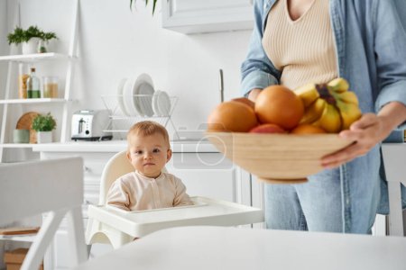 little kid in baby chair looking at camera near mom with bowl of fresh fruits on blurred foreground